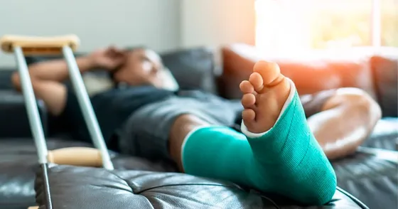 Pessoa com a perna imobilizada por gesso, deitada no sofá ao lado de uma muleta